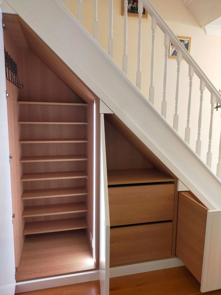 Side view of a stair case featuring two open cupboard doors showing shelving and hanging space in the taller one, and two drawers with space above in the one to its right.