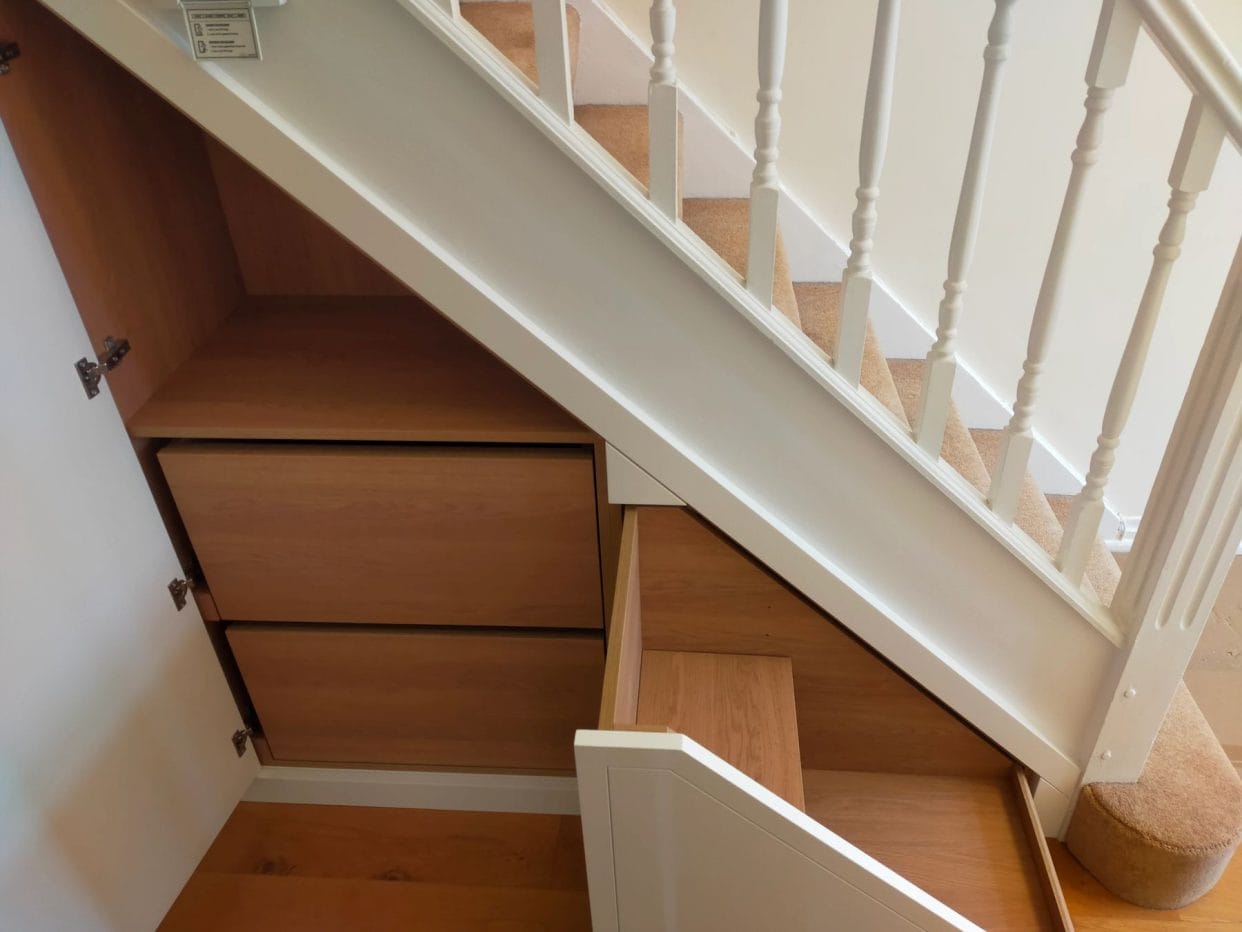 Side view of a stair case featuring two open cupboard doors showing two drawers with space above in the left one, the right one features innovative pull-out shoe storage in a stepped design.