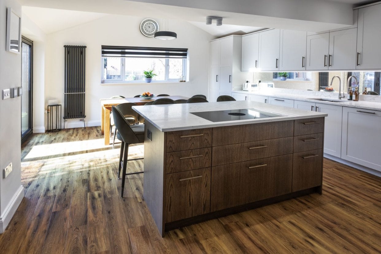 A modern kitchen featuring rich wood flooring, a central island with umber-toned panelling, and off-white shaker kitchen cabinets. There is a dining table at the far end under the window.