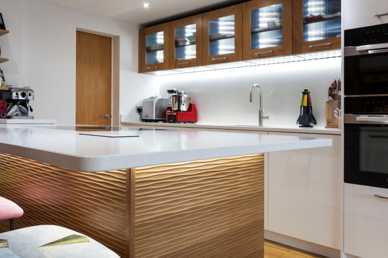 A mind-century inspired kitchen with gloss white acrylic cabinetry combined with oak, glass fronted eye-level cabinets, along with a statement kitchen island with wave-design panelling. 