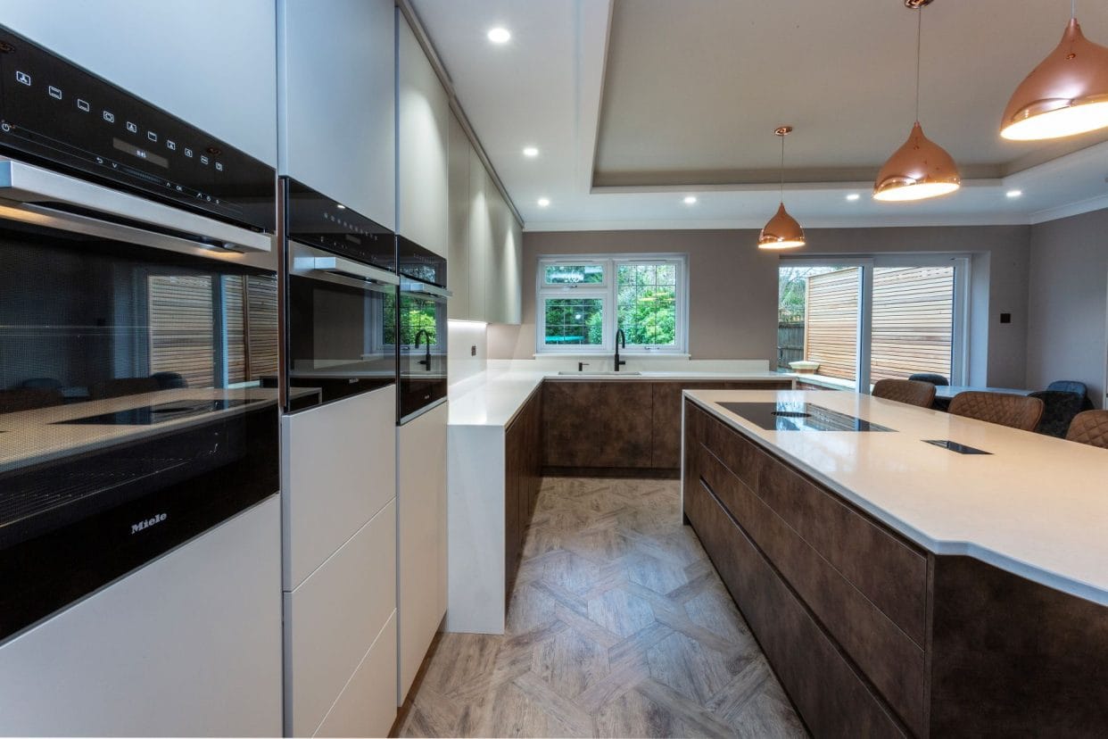 A contemporary kitchen with simple flat panel doors and a large feature island with laminated panels in a rich mottled bronze tone. Copper pendant lights hang over the island and there is seating on the other side. 