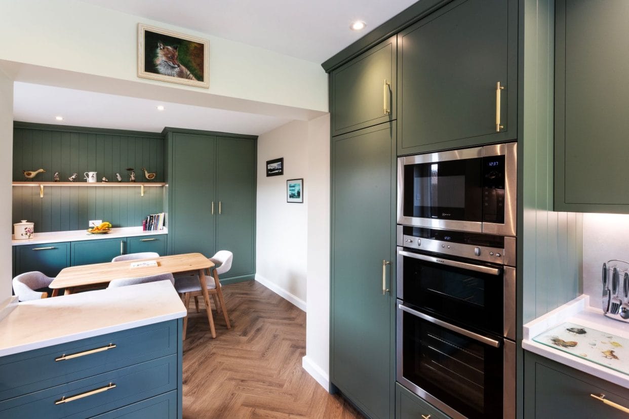  kitchen painted in a rich teal green with brass bar handles and a rich parquet floor. There is tongue and groove feature panelling on the far wall. 