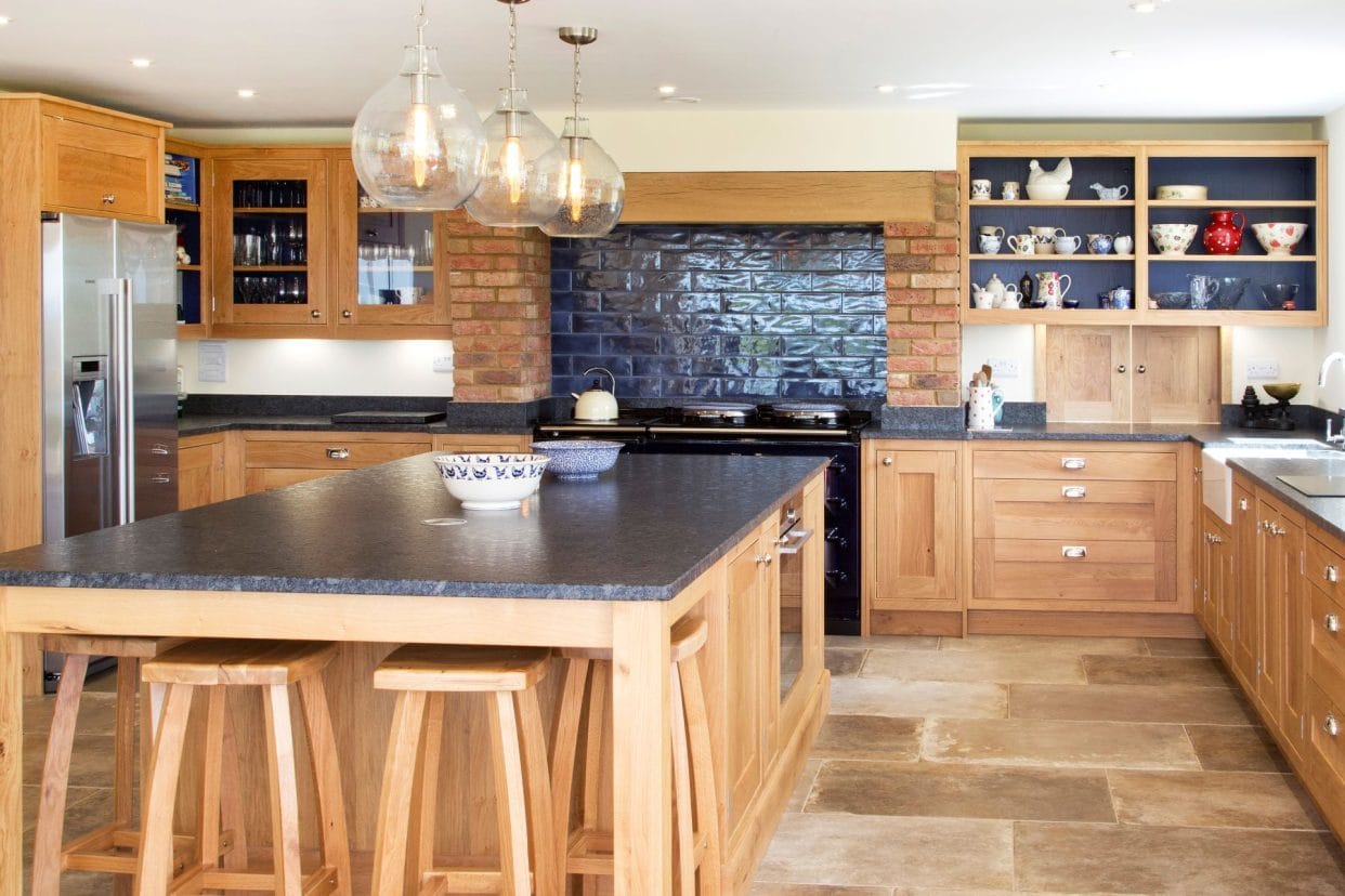 A traditional kitchen with oak cabinetry, exposed brick and flagstone floor. There is a generous central island with stool seating and grey granite countertop. 