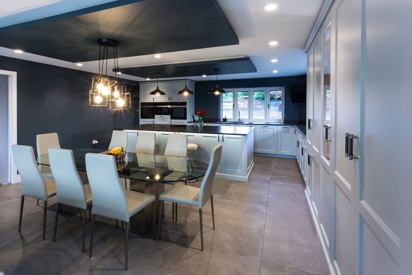 A modern shaker style kitchen with light cabinetry on one side and charcoal feature walls around the kitchen window and alongside an intimate dining area, with dark dropped ceiling panels for pendant lights.