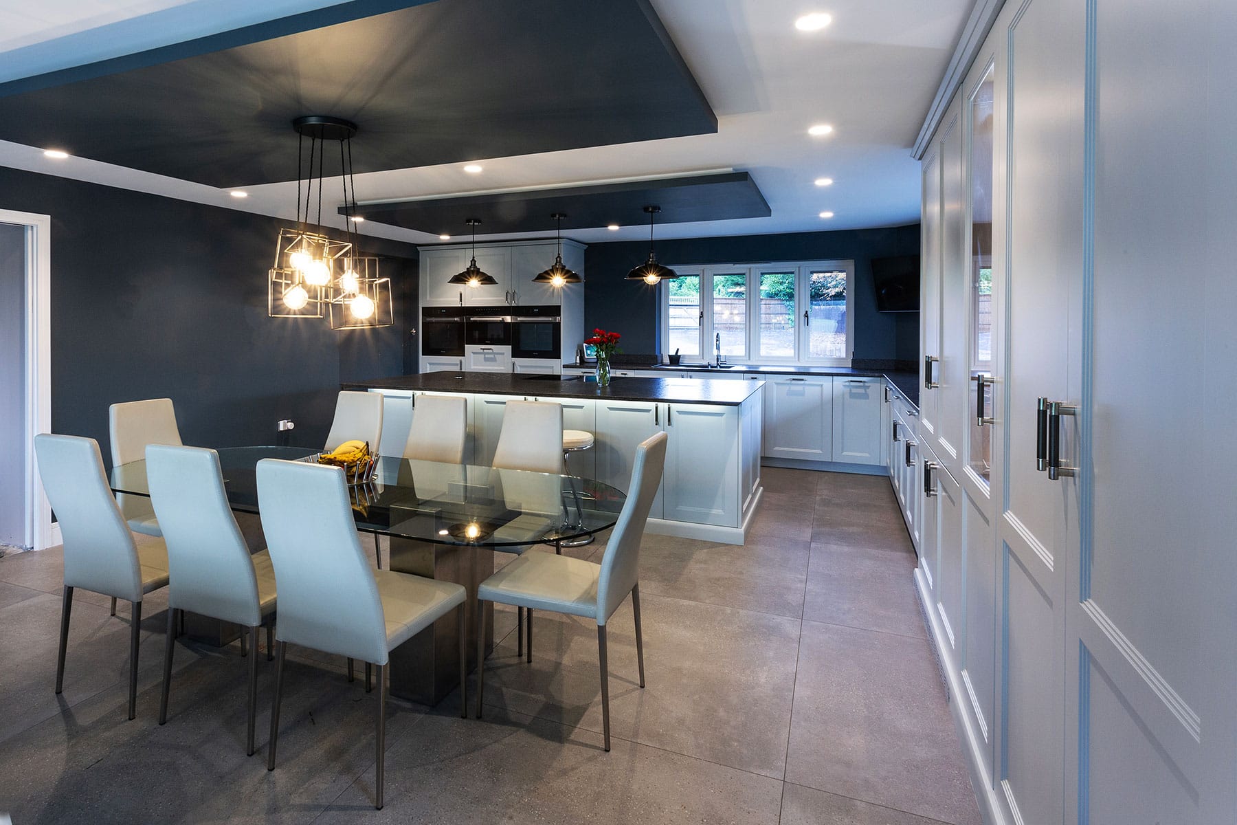 A modern shaker style kitchen with light cabinetry on one side and charcoal feature walls around the kitchen window and alongside an intimate dining area, with dark dropped ceiling panels for pendant lights.