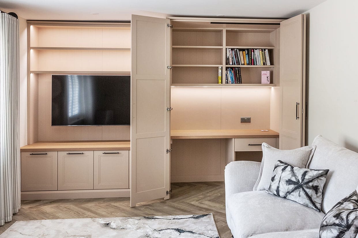 A bank of cabinetry painted in a soft peach – on one side there is a media unit, on the other a study area concealed by bi-fold doors. There is also a sofa in this multi-functional space.
