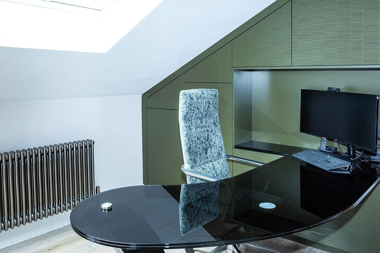 A study in a loft space with sloping ceiling. The flat panel cabinetry is a rich sage green, a curved black glass desk has a computer screen on top and a grey office chair.