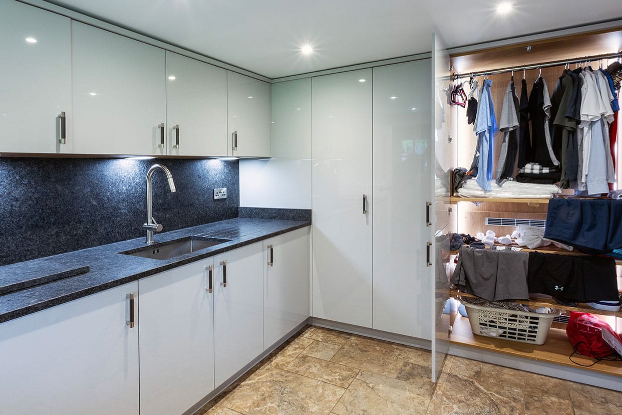 A spacious utility room with light grey gloss acrylic floor-to-ceiling cabinetry and split-level cabinets with a sink unit. Double doors are open to reveal neatly handing laundry with internal lighting.