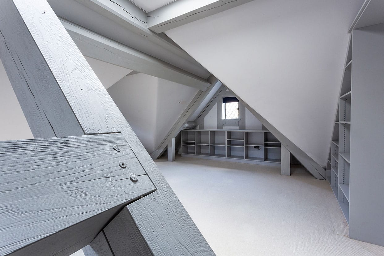 A loft space with sloping ceilings on either side. Bespoke shelving has been created to fit the wall space and everything is painted in a light grey tone.