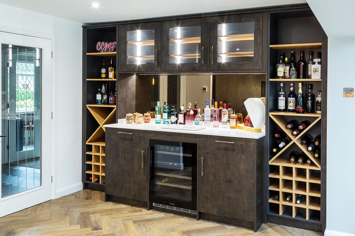 A home bar along the entire wall of a kitchen with a central wine fridge below a white quartz bar top. The surrounding cabinetry is dark brown with oak wine racks.