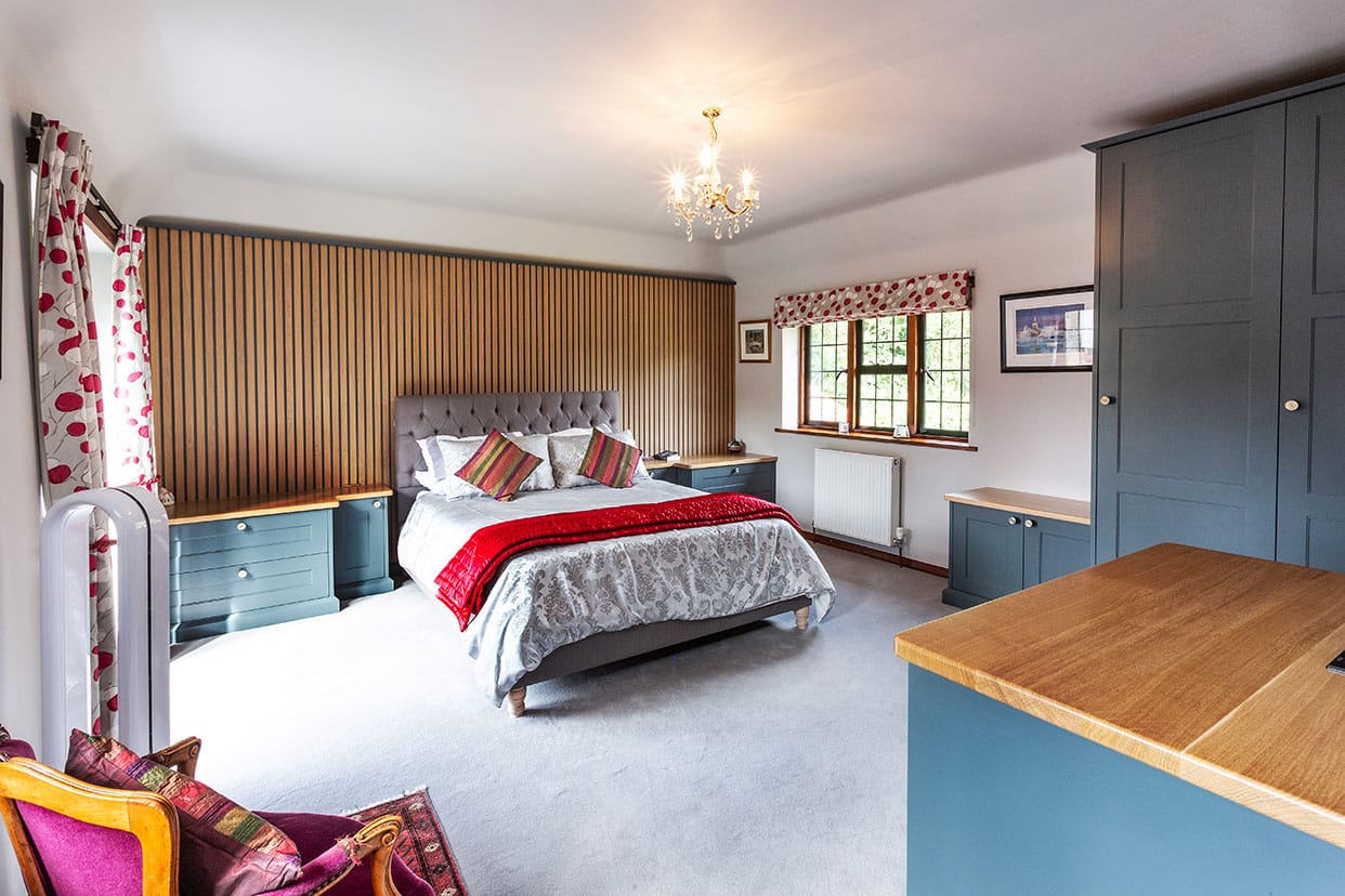 A spacious bedroom with blue painted cabinetry combined with oak. Behind the bed is a wood panelled feature wall.
