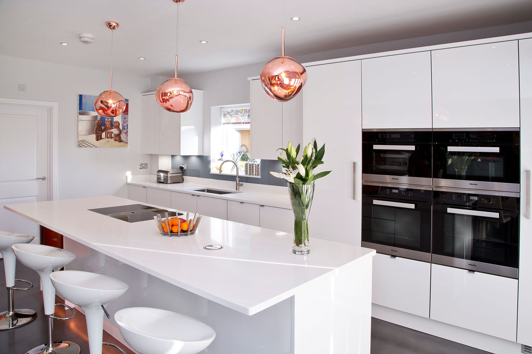 A modern kitchen with a high gloss white theme throughout the acrylic cabinetry and crisp white quartz for the kitchen island, above which are copper pendant lights.