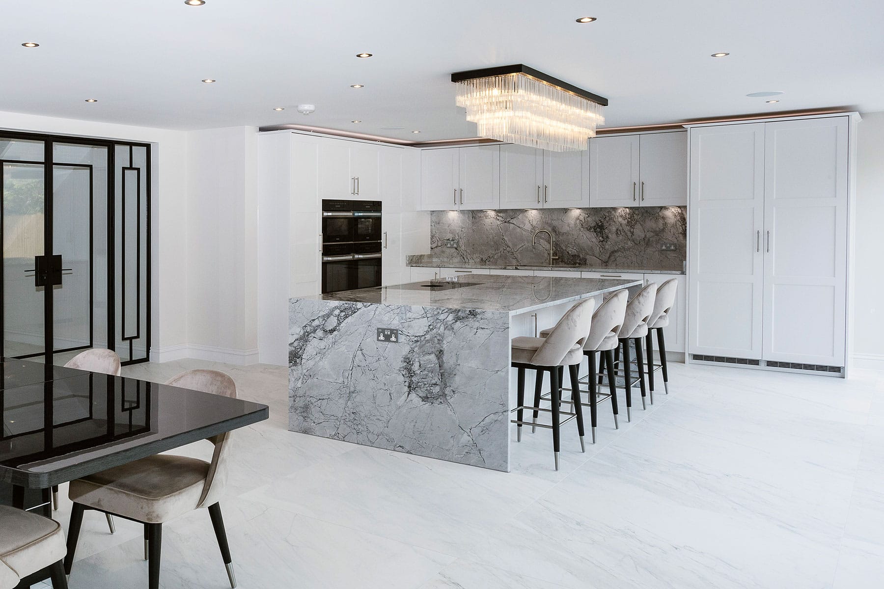 A spacious kitchen diner with a grey marbled theme throughout the flooring and kitchen island’s waterfall-style countertop. The cabinetry is white-painted shaker style. There are black accents in the furniture and crittall-style internal door.