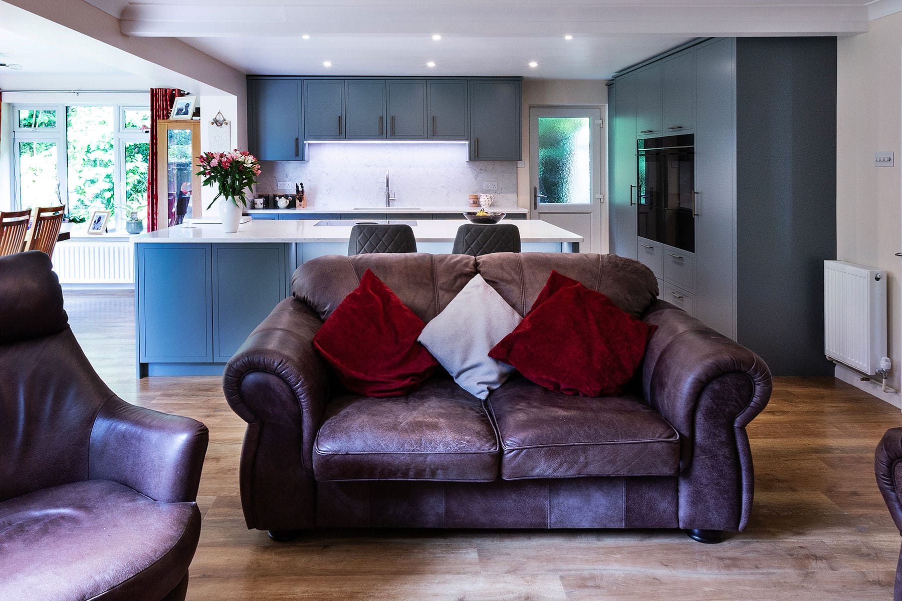 A kitchen and living space with teal painted cabinetry and rich wooden floor that continues into a comfortable seating area with aged brown leather sofa and chair.