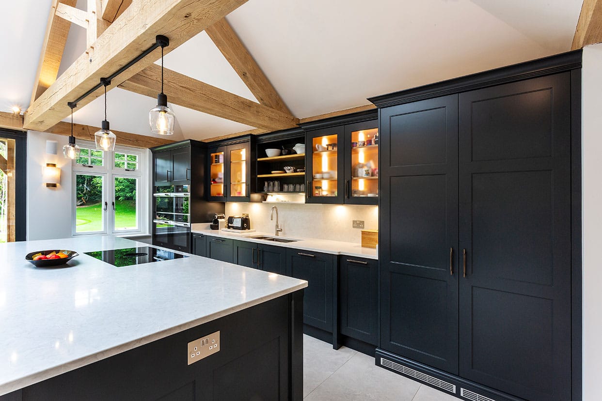 A modern shaker kitchen with vaulted oak-beamed ceiling. The cabinetry is painted in black with the contrast of crisp white quartz countertops.