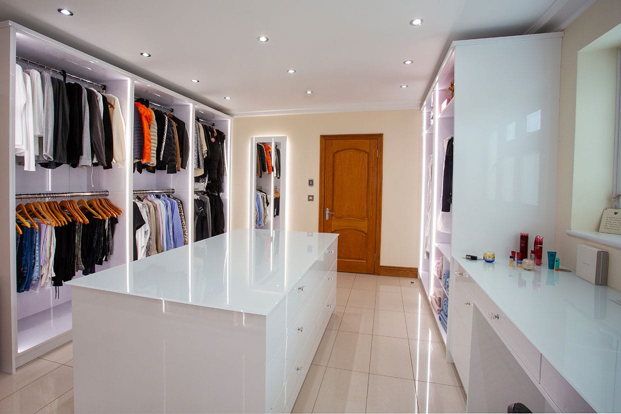 A spacious dressing room with white gloss cabinetry and cream polished floor tiles. The wardrobes are open to reveal neatly hanging clothes, there is a large, central chest of drawers.