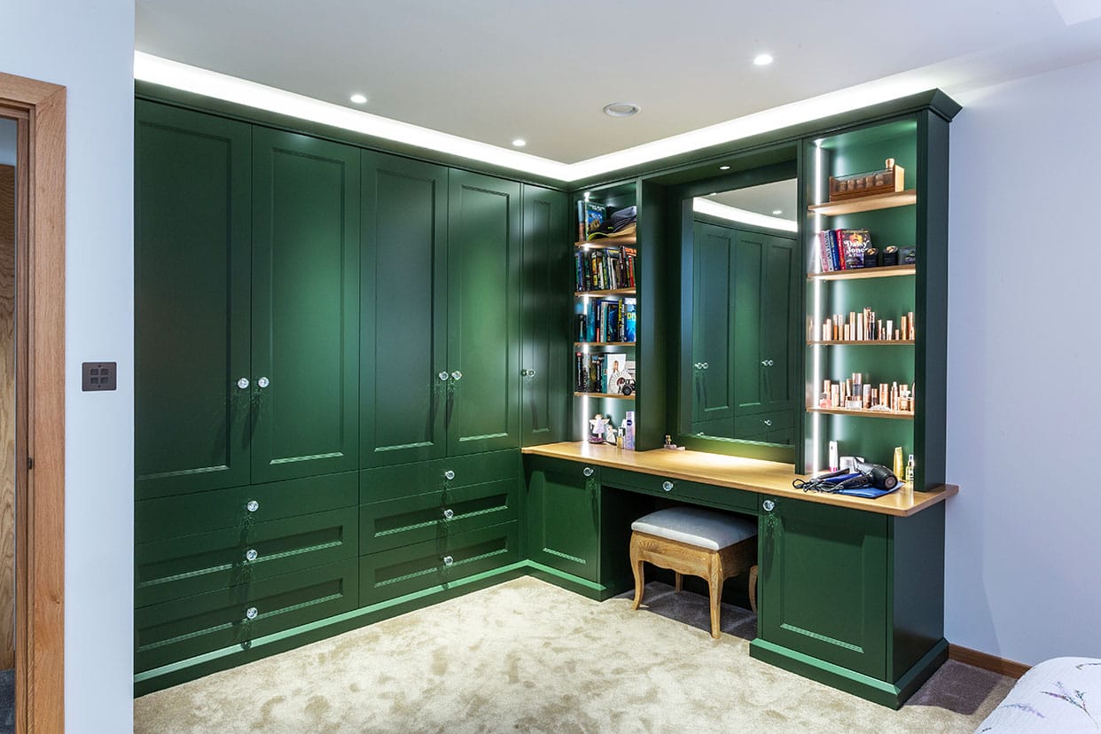 The corner of a bedroom featuring L-shaped wardrobe and dressing table cabinetry painted in a rich fir green.