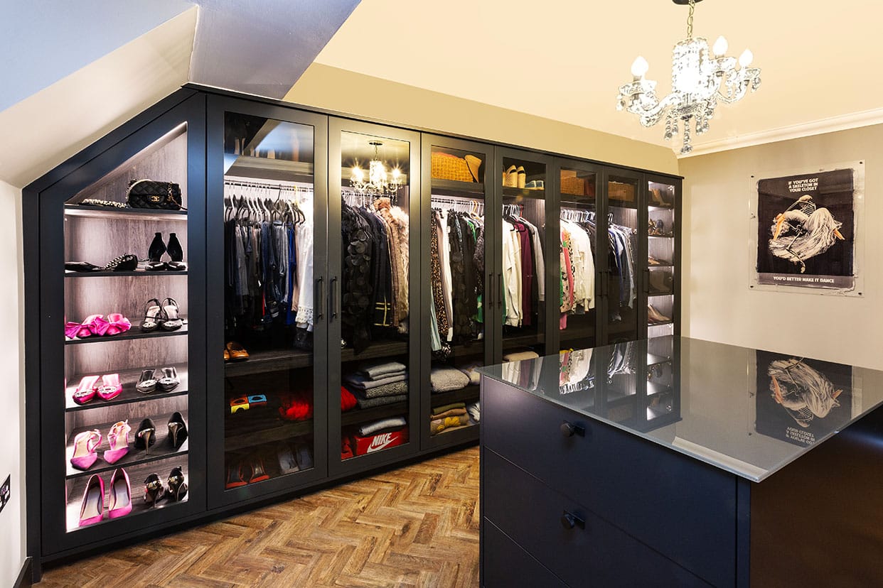 A Dressing room with black floor-to-ceiling wardrobes and chest of drawers. The cabinetry features glass doors so the clothes hanging within are visible. There is wooden parquet flooring along with a feature chandelier.