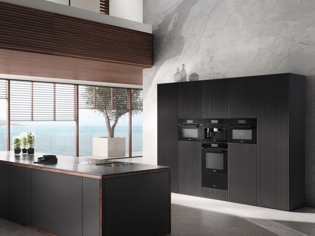 A beautiful, minimalist kitchen with dark wood tones. There is a bank of Miele ovens set into dark fluted cabinetry.