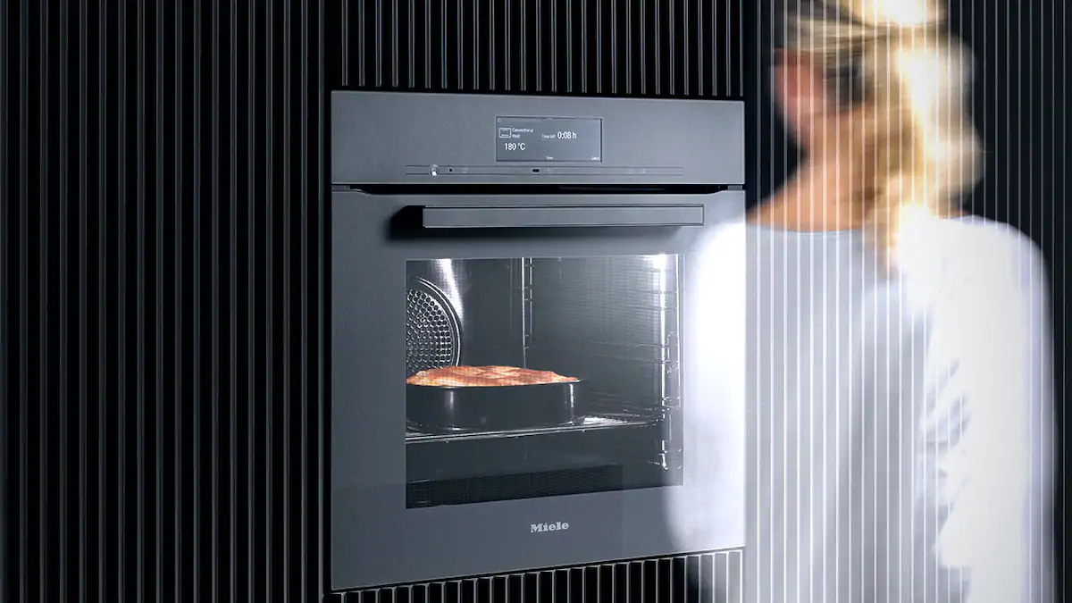 The impression of a woman walking past the Miele steam combi oven, inside a large cake is slowly rising and has a light brown top.
