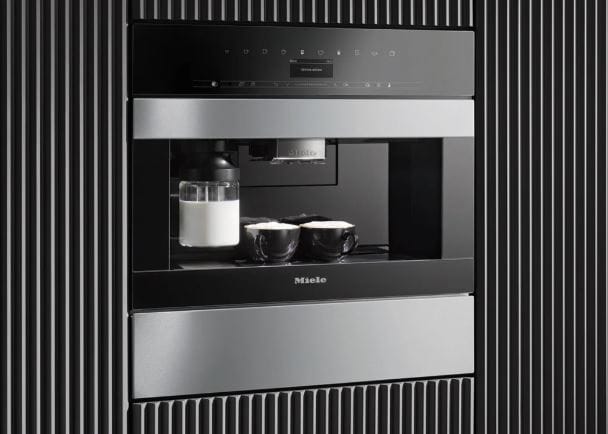 A sleek Miele built-in coffee machine with two cups topped with frothed milk alongside a glass milk jug.