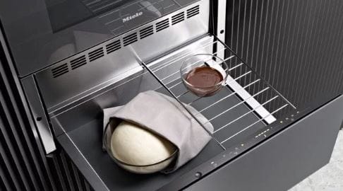 An open Miele warming drawer with dough in a glass bowl alongside a glass bowl of chocolate sauce.