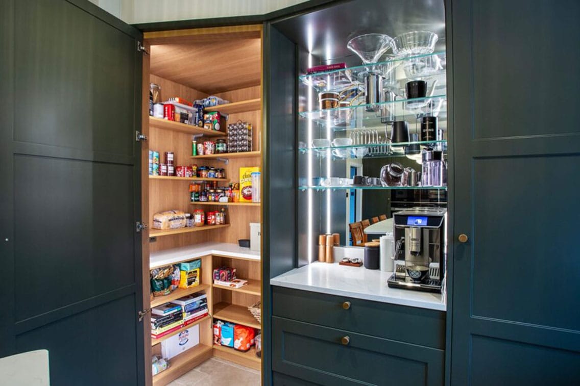 A stylish kitchen in a deep teal-painted shaker style, there is an open door to a well-organised pantry cupboard alongside a bespoke bar with glass shelving and mirror splashback.