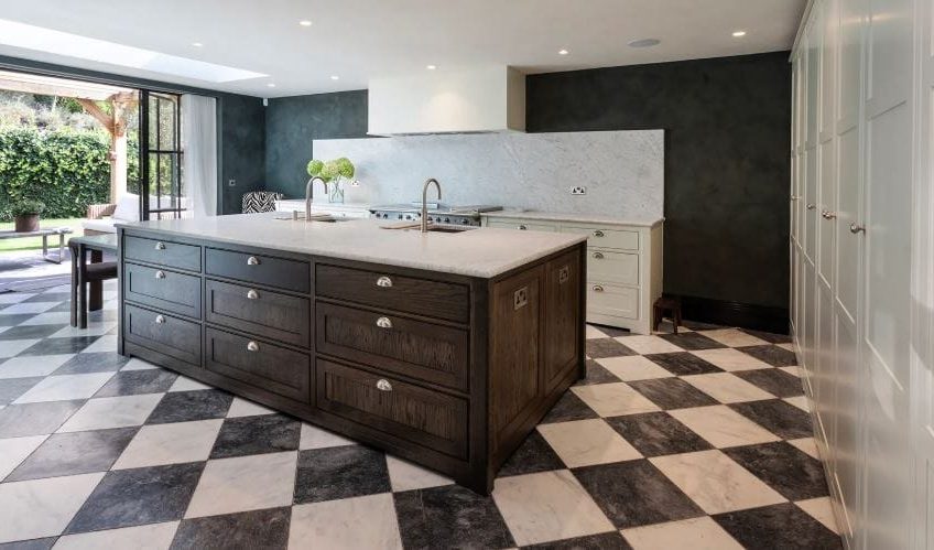 A striking open plan kitchen with a large island in traditional shaker style, there is a full-height wall of cabinetry to the right and a marble-backed dresser-style bank to the back of the room housing the oven. The floor is a stunning black and white tile design.