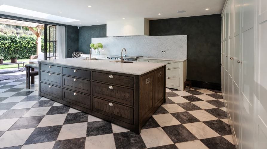 A striking open plan kitchen with a large island in traditional shaker style, there is a full-height wall of cabinetry to the right and a marble-backed dresser-style bank to the back of the room housing the oven. The floor is a stunning black and white tile design.
