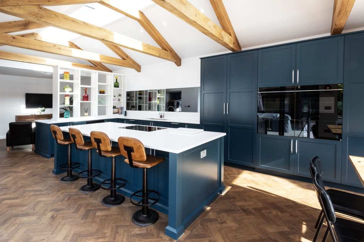 A rich blue painted modern shaker kitchen with vaulted oak beams and simple white paintwork and countertops.