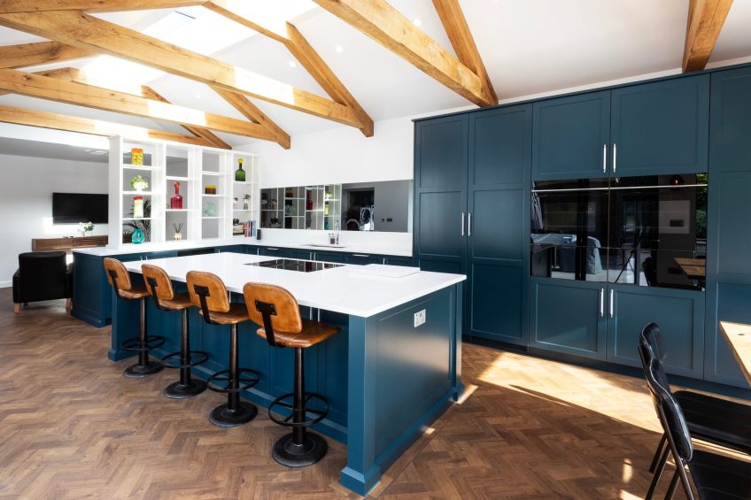 A rich blue painted modern shaker kitchen with vaulted oak beams and simple white paintwork and countertops.