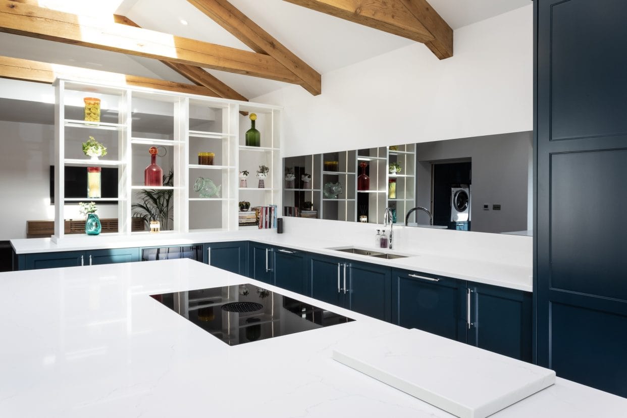 An open-plan kitchen with white open shelving to one end, with a large kitchen island finished in pure white quartz. A BORA X Pure hob is flush-set into the centre.