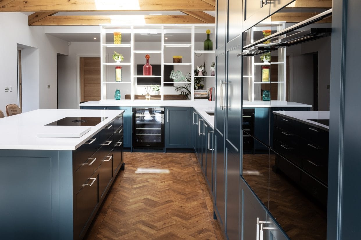 An open-plan kitchen with white open shelving to one end, featuring an island to the left and full height cabinetry opposite with built-in Siemens appliances.