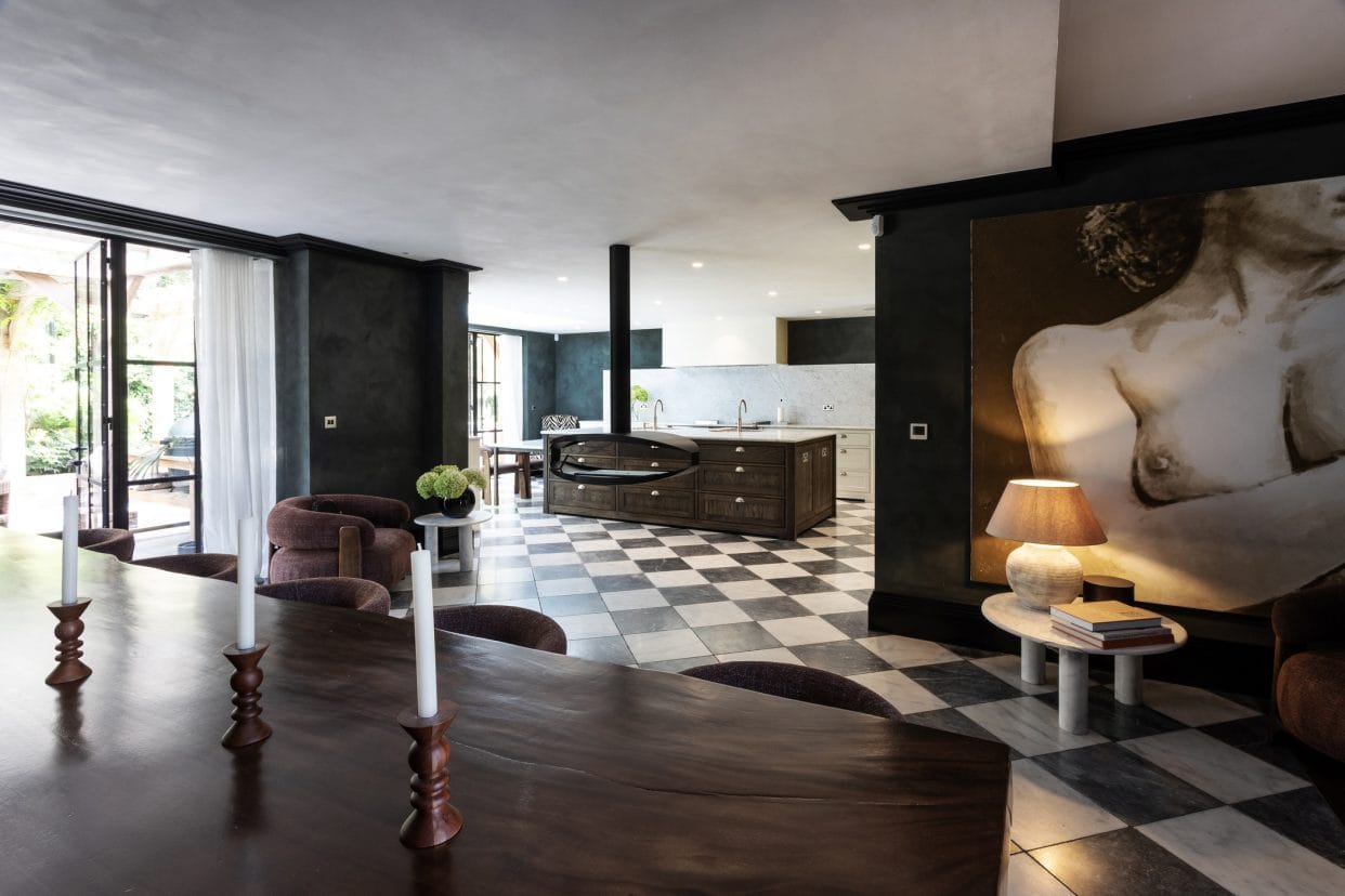 View towards a striking modern shaker kitchen island in deep rich wood tones with marble countertop, there are black and white Revival-style floor tiles connecting the space to the dining area with a large mahogany dining table and rich brown upholstered chairs.