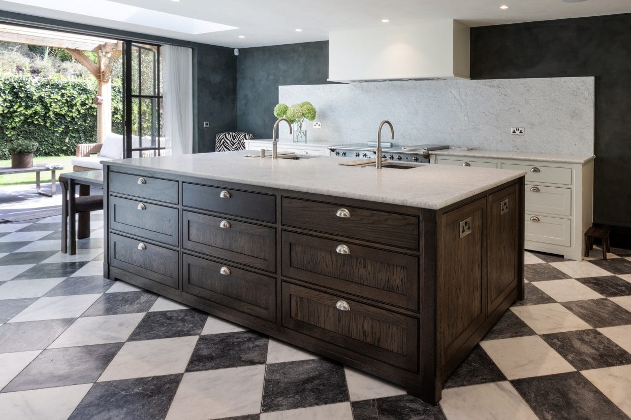 A striking modern shaker kitchen island in deep rich wood tones with marble countertop, there are black and white Revival-style floor tiles, a crittall-style patio door opens onto the garden