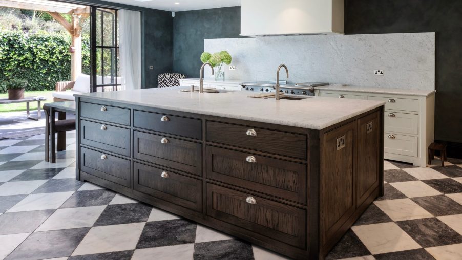 A striking modern shaker kitchen island in deep rich wood tones with marble countertop, there are black and white Revival-style floor tiles, a crittall-style patio door opens onto the garden