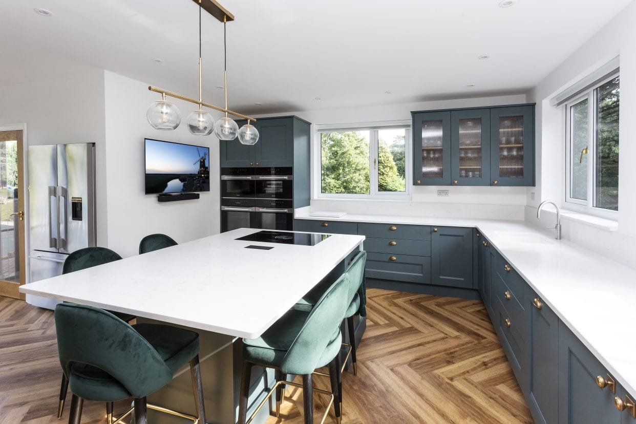 A traditional style shaker L-shaped kitchen finished in a deep turquoise with spacious areas of white quartz work surfaces, a combination of full height, waist height and split-level cabinetry, and a generous central island with plenty of seating.