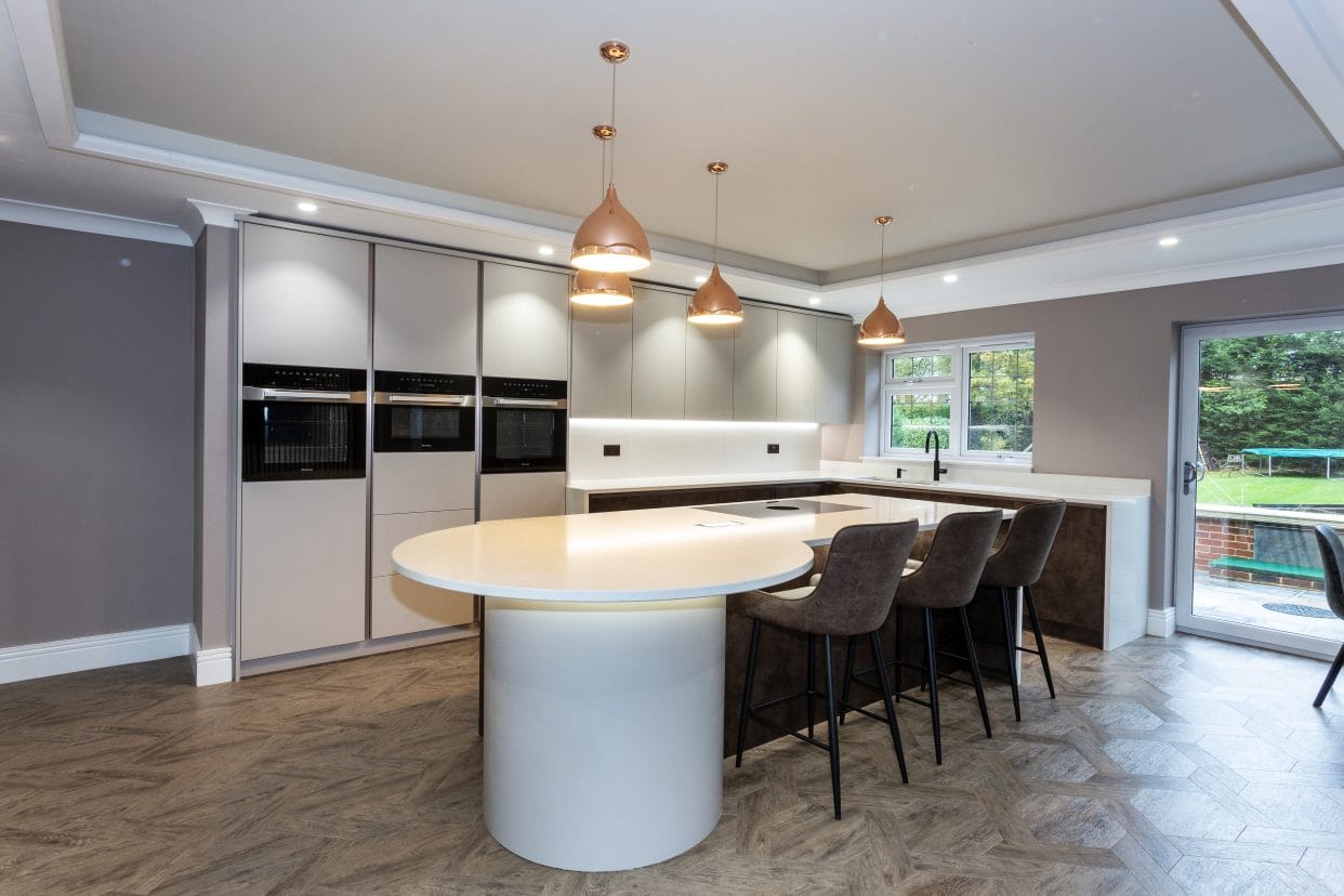 A modern kitchen with flat panel doors with an L-shaped layout extending under a kitchen window. A bespoke kitchen island with drum base at one end and integrated seating separates the kitchen from the living area.
