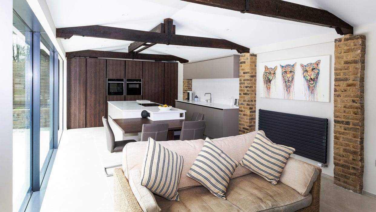 A modern L-shaped kitchen at one end of a large open-plan kitchen/living room. There is understated flat panel cabinetry in natural tones to complement exposed beams and brickwork