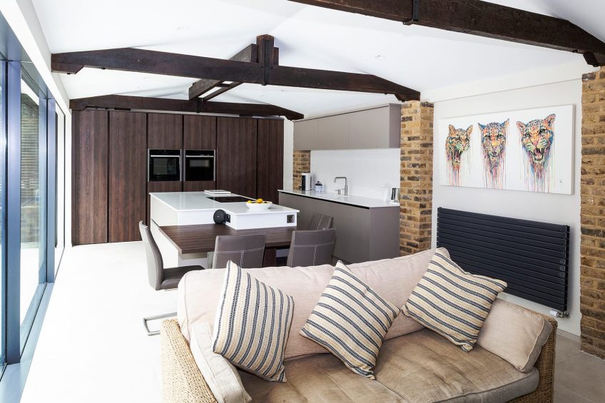 A modern L-shaped kitchen at one end of a large open-plan kitchen/living room. There is understated flat panel cabinetry in natural tones to complement exposed beams and brickwork